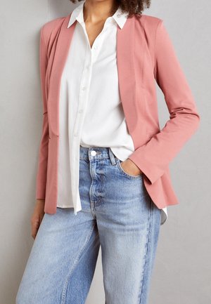 Kobieta w jasnoróżowej marynarce na białej koszuli zapinanej na guziki i jasnoniebieskich, wysokich dżinsach, z jedną ręką w kieszeni, na szarym tle.