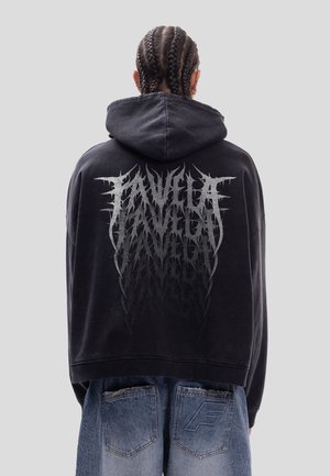 Persona con el cabello trenzado que lleva una sudadera negra con capucha con un gran texto abstracto plateado en la espalda y pantalones vaqueros azules y sueltos.