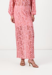 Jupe longue en dentelle rose avec motif floral et superposition transparente, associée à des sandales à talons hauts beiges à lanières. Texture lisse, design ajusté.