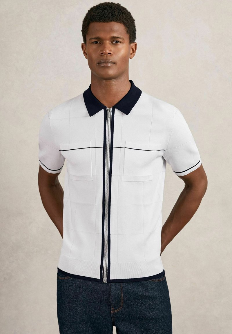 Modelo masculino vistiendo una camisa blanca de manga corta con un cuello azul marino y cremallera en la parte delantera, de pie contra un fondo neutral, con las manos detrás de la espalda.