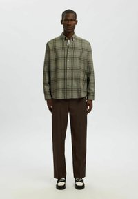 Zelena karirana srajca s sproščenim krojem, v kombinaciji z rjavimi hlačami. Kratki rokavi in ukrivljen rob z gladko teksturo.