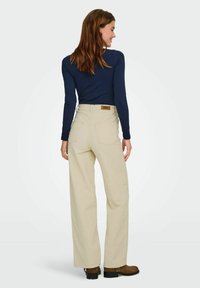 Pantalons beige taille haute à jambes larges avec une finition texturée, dotés de poches arrière et d'un patch logo marron au-dessus de la taille.