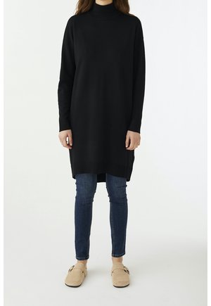 Abito tunica a collo alto in maglia nera con maniche lunghe, che si estende sotto i fianchi. Abbinato a jeans in denim blu e scarpe slip-on beige.