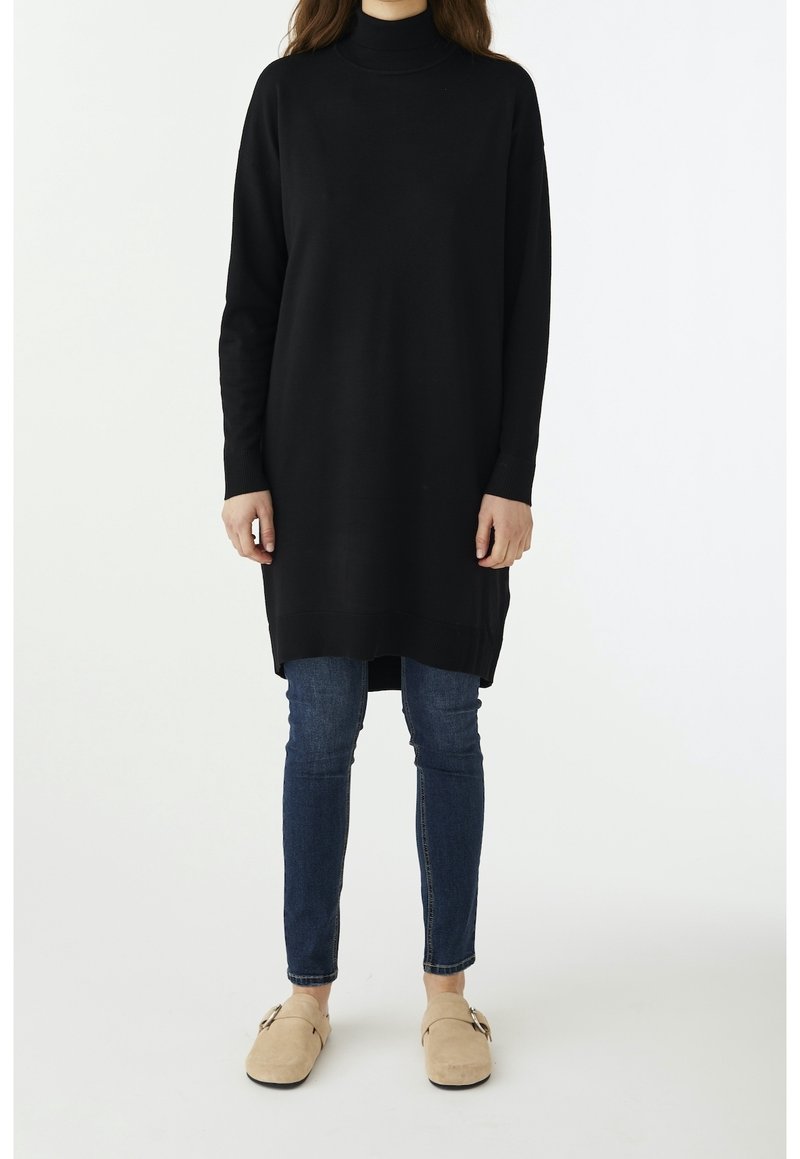 Abito tunica a collo alto in maglia nera con maniche lunghe, che si estende sotto i fianchi. Abbinato a jeans in denim blu e scarpe slip-on beige.