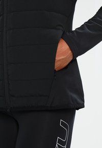 Svart väst med quiltat mönster, som har en dragkedjeförsedd sidoficka och en slät, välsittande silhuett, tillverkad av elastisk tyg.