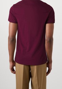 Krátký rukáv tričko z burgundské bavlny, s kulatým výstřihem a volným střihem, zobrazené zezadu, v kombinaci s béžovými kalhotami.