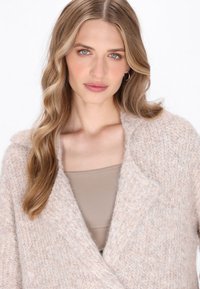Una donna bionda con capelli ondulati indossa un cardigan beige a maglia testurizzata sopra una maglietta taupe, posando su uno sfondo bianco uniforme.