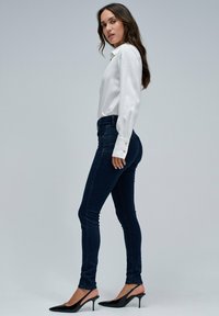 Kvinna i vit långärmad blus, mörka slim-fit jeans och svarta högklackade slingback-skor som står sidledes mot en grå bakgrund.