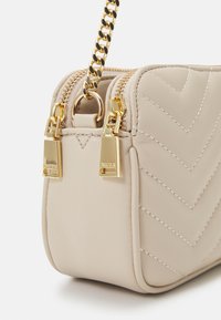 Beige faux skinnveske med chevron-søm, to gullglidere og en gullkjede stropp. Rektangulær form med en jevn tekstur.