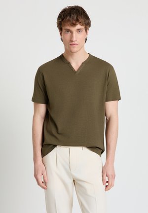 Giovane uomo che indossa una maglia henley a maniche corte verde oliva e pantaloni color crema, in piedi contro uno sfondo chiaro e uniforme.