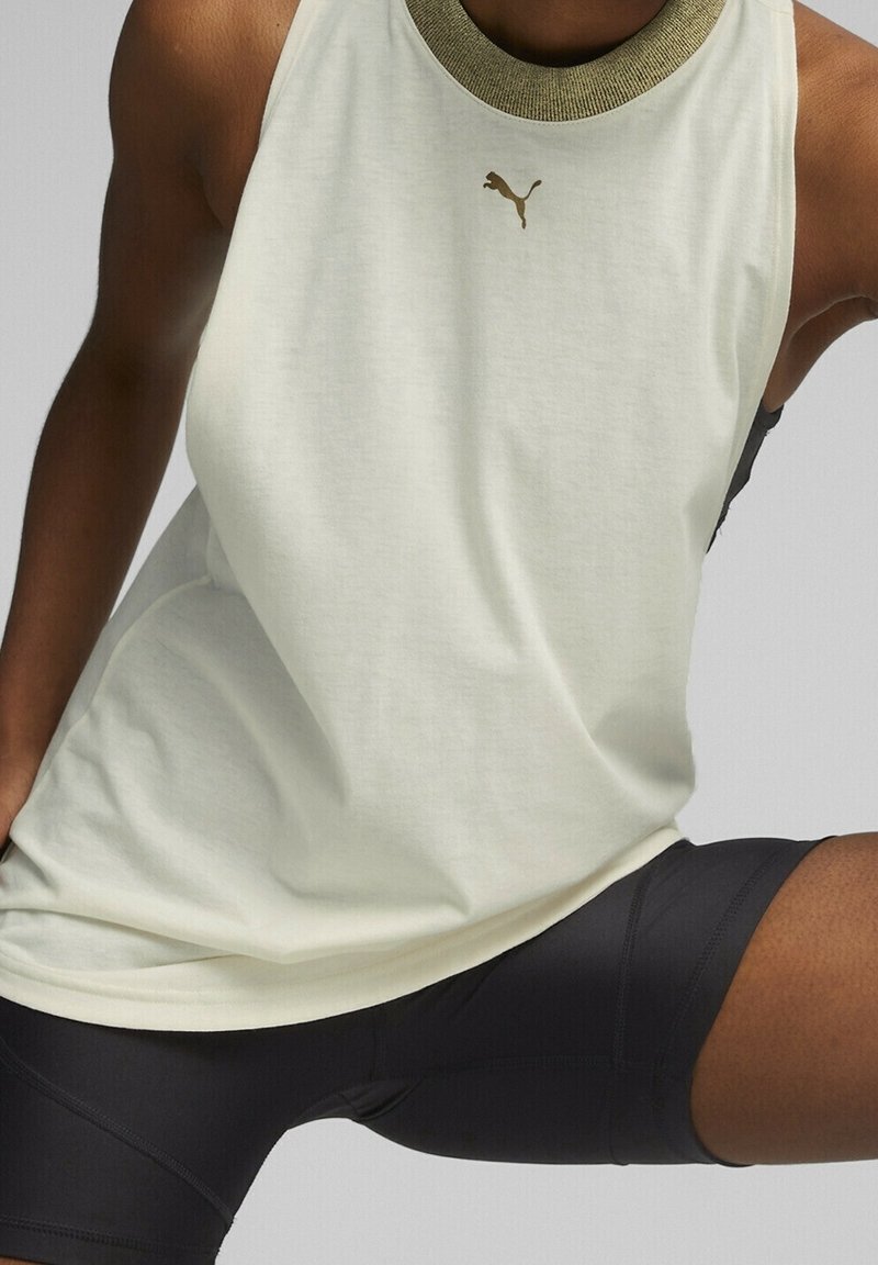 Light beige sleeveless athletic top with a textured dark green collar, featuring a small gold logo. Paired with black fitted shorts.