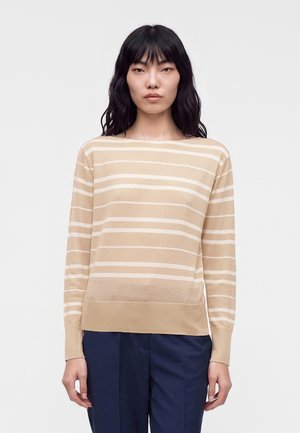 Femme aux cheveux foncés portant un pull à manches longues beige et blanc à rayures et un pantalon bleu foncé, debout devant un fond clair uni.