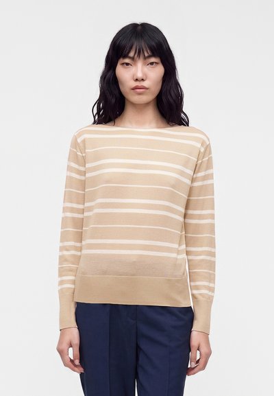 Femme aux cheveux foncés portant un pull à manches longues beige et blanc à rayures et un pantalon bleu foncé, debout devant un fond clair uni.