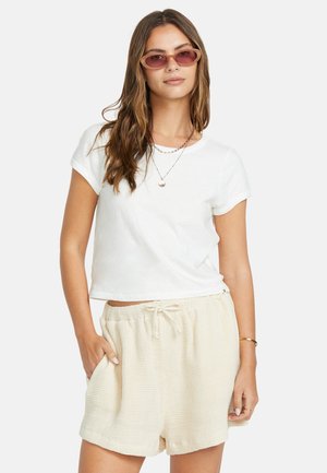 T-shirt blanc court à manches courtes, associé à un short beige texturé. Le mannequin porte des lunettes de soleil roses et des colliers superposés.