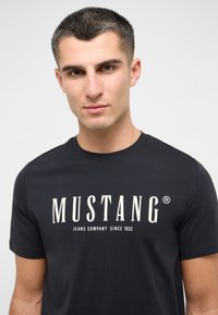 Melna kokvilnas T-kreklu ar lielu baltu "MUSTANG" logo un tekstu "JEANS COMPANY SINCE 1932", īsām piedurknēm un apaļu kakla izgriezumu.