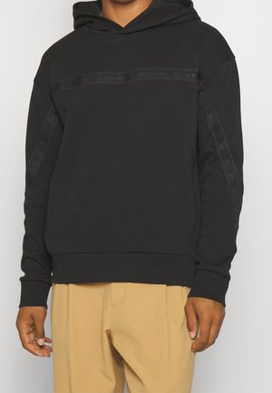 Person trägt einen schwarzen Calvin Klein Hoodie mit gebrandetem Klebeband auf Brust und Ärmeln, kombiniert mit beigen Hosen, steht vor einem schlichten Hintergrund.