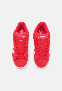 Zapatillas de ante rojo con acentos blancos, cordones planos y puntera redondeada. El interior cuenta con un forro rojo en contraste y un detalle del logotipo.
