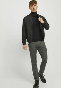 Crna bomber jakna preko crnog turtleneck pulovera, uparena sa sivim slim-fit hlačama i crnim cipelama na vezivanje. Jednostavan, moderan stil.