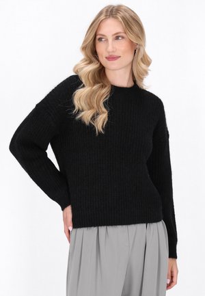 Zwarte gebreide trui met een losse pasvorm, lange mouwen en ribbelstructuur. Draagend over lichtgrijze geplooide broek, met een ronde halslijn.