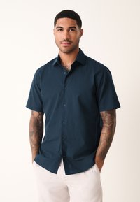 Korte mouwen, donkerblauw overhemd met knopen gemaakt van textuurstof, met standaard kraag en lichtblauwe knopen, gedragen met lichte broek.