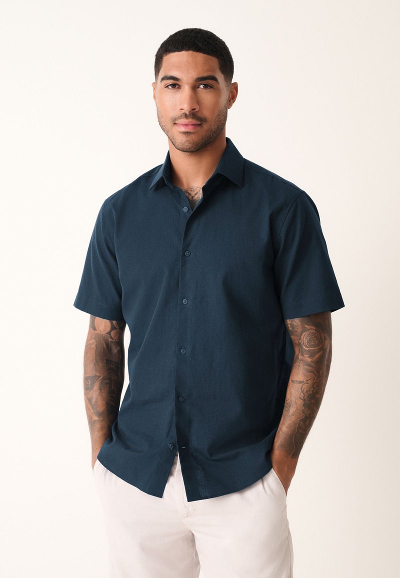 Korte mouwen, donkerblauw overhemd met knopen gemaakt van textuurstof, met standaard kraag en lichtblauwe knopen, gedragen met lichte broek.