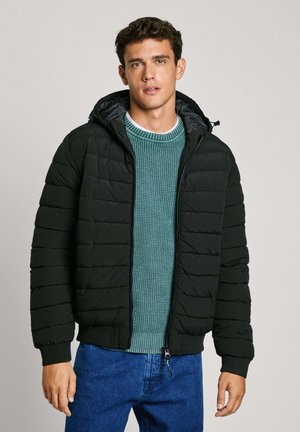 Chaqueta acolchada negra con costuras horizontales, capucha y cremallera frontal, llevada sobre un suéter de punto verde y jeans de mezclilla azules.