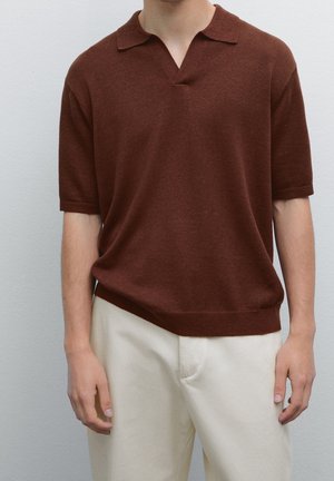 Personne portant un polo en maille à manches courtes marron et un pantalon blanc cassé, debout devant un fond gris clair uni.