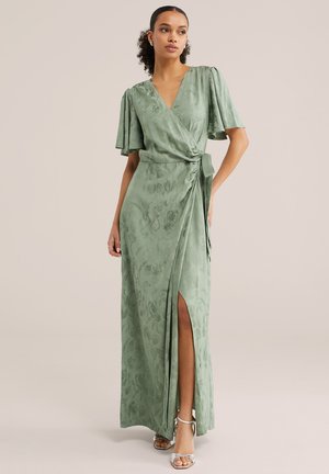 Vrouw draagt een groene bloemenwrapjurk met zwierige mouwen en een hoge split tot aan de dij, gecombineerd met zilveren sandalen met hoge hakken, staande tegen een beige achtergrond.