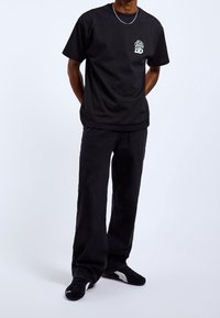 Czarny t-shirt z krótkimi rękawami, z małą kolorową grafiką tygrysa na klatce piersiowej, zestawiony z czarnymi szerokimi spodniami i czarnymi trampkami.