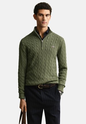 Junger Mann trägt einen olivgrünen, kabelgestrickten Halb-Reißverschluss-Pullover über einem dunklen Hemd und hält mit einer Hand eine braune Ledertasche.