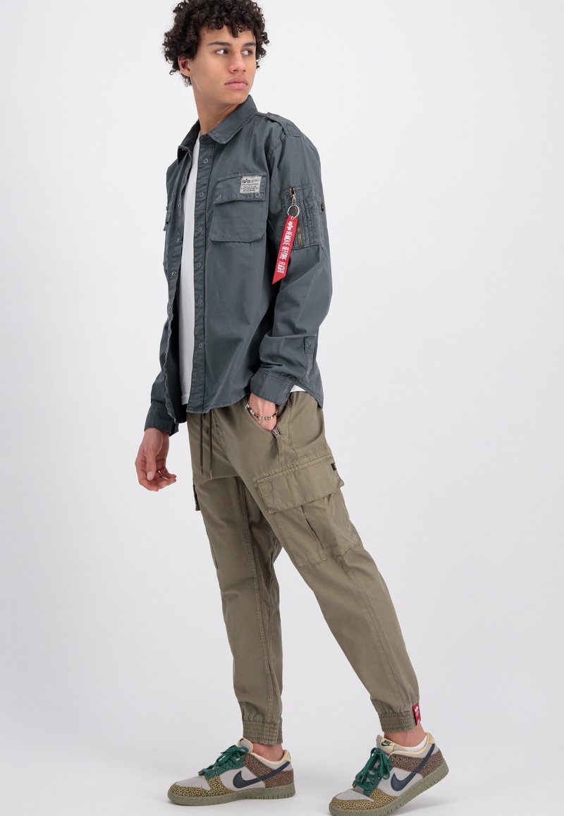 Giovane uomo che guarda di lato indossando una camicia a bottoni grigio scuro con un'etichetta rossa, pantaloni cargo beige e sneakers Nike multicolori.