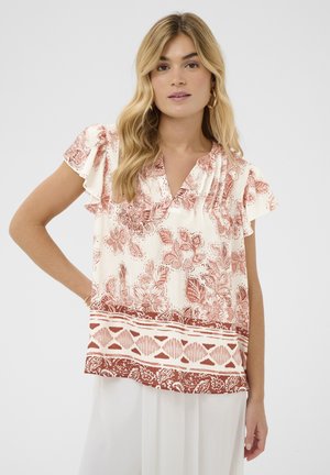 Blonde vrouw die een witte blouse draagt met roestkleurige bloemen- en geometrische patronen, korte losvallende mouwen en een witte broek, staand tegen een effen achtergrond.