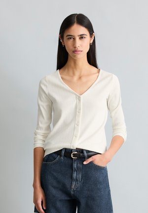 Frau mit langen dunklen Haaren trägt ein cremefarbenes Oberteil mit Knöpfen und dunkelblaue Jeans mit schwarzem Gürtel und steht vor einem hellgrauen Hintergrund.