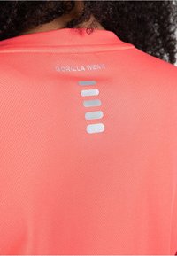 Lichtgewicht, heldere koraalkleurige sportshirt met gaastextuur. Bevat een zilveren "GORILLA WEAR" logo en verticale gestreepte accenten op de achterkant.