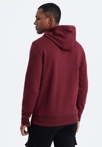 Felpa bordeaux realizzata in tessuto morbido, con cappuccio regolabile e polsini e fondo a costine. Il modello è mostrato di spalle, con pantaloni neri.