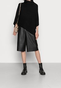 Femme portant un poncho noir côtelé, un short en cuir noir jusqu'aux genoux, des bottines noires à la cheville, et un petit sac bandoulière beige à imprimé serpent.