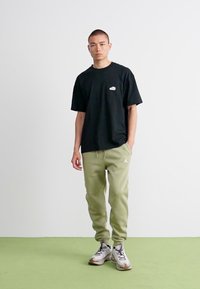 Mand stående imod en hvid væg iført sort t-shirt, lysegrønne sweatpants med logo og grå sneakers på grønne gulv.