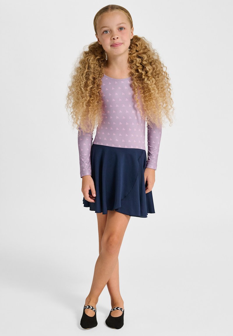 Młoda dziewczyna z długimi kręconymi włosami, ubrana w lawendowy top z długim rękawem, granatową spódnicę i czarne baletki, stojąca ze skrzyżowanymi nogami.