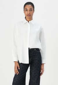 Weißes Hemd mit Knöpfen und einem kleinen Herzaufnäher, kombiniert mit dunkel gemusterten Jeans mit Schlaufen. Der Stoff wirkt glatt und leicht.