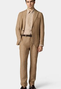 Completo in lino di un beige chiaro con una vestibilità rilassata, dotato di una giacca con un solo bottone, pantaloni coordinati e una camicia a righe sotto.