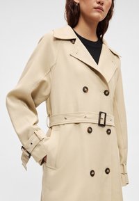 Μπεζ διπλόμπουτο καμπαρντίνα με μεγάλα καφέ κουμπιά, ζώνη στη μέση και φαρδιές πέτες. Διαθέτει θηλιές για ζώνη και λεπτομέρειες στα μανίκια.