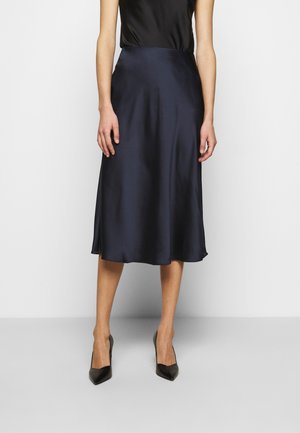 Vrouw draagt een zwarte satijnen midirok gecombineerd met een mouwloze zwarte top en zwarte puntige hakken, staand tegen een effen achtergrond.