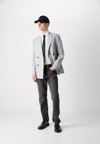 Gray checked double-breasted blazer over white shirt and black tie, paired with dark gray jeans and black shoes, topped with a black cap.