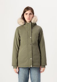 Olive roheline parka jakk vale-karvakaelusega, nööbiklapp, küljetaskud ja vabalt langev lõige, kantud siniste teksapükste peal.