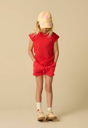 Jong kind in een rood mouwloos topje en korte broek, met een gele pet met roze bloemen, witte sokken en beige sneakers, staand met de handen in de zakken.