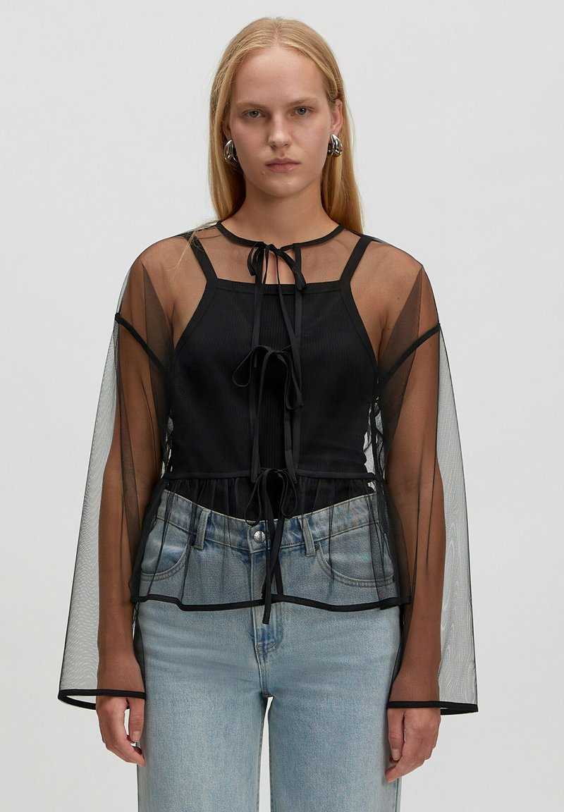 Junge Frau, die ein durchsichtiges schwarzes Langarmoberteil mit Bändern über einem schwarzen Tanktop und hellblauen hochgeschnittenen Jeans trägt, steht vor einem schlichten Hintergrund.