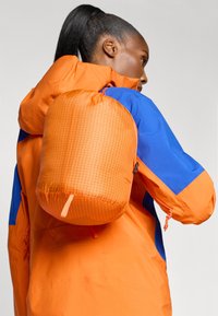 Rucsac portabil portocalie cu cordon, cu un model în grilă, cu o suprafață texturată. Purtați pe spate peste o jachetă strălucitor portocalie și albastră.