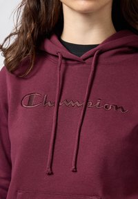 Sudadera color burdeos hecha de tejido de sudadera, con capucha ajustable y logo bordado de "Champion" en el pecho.
