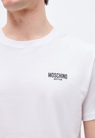 Άνδρας με λευκό t-shirt Moschino Active, που δείχνει το πάνω μέρος του στήθους και το λαιμόκοψη με μαύρο λογότυπο στο αριστερό μέρος.