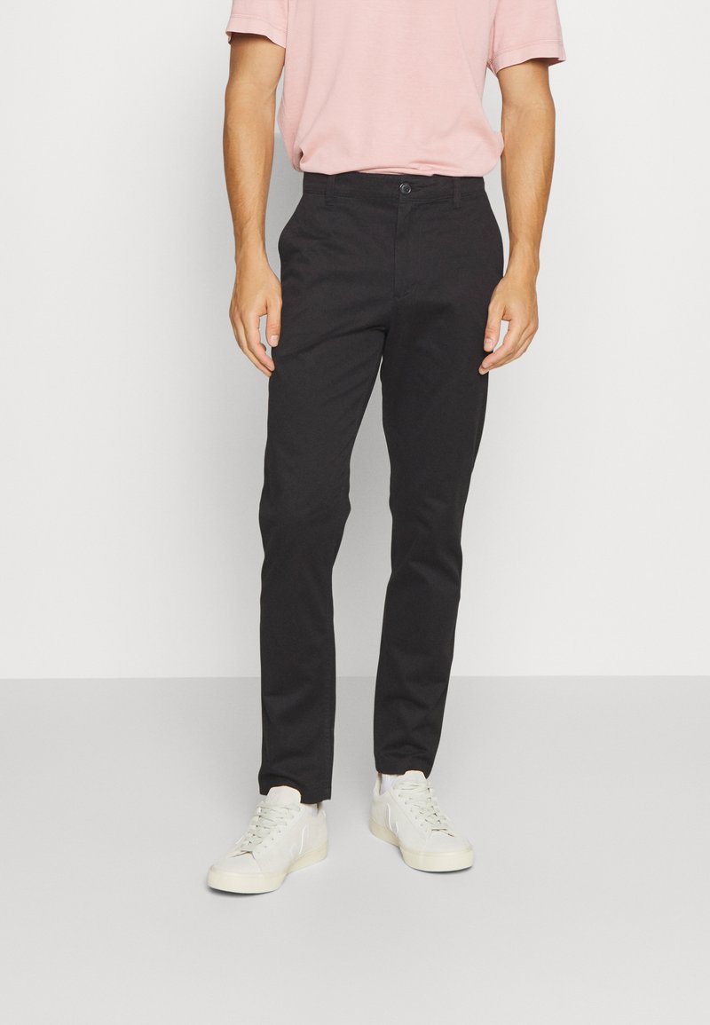 Pantaloni neri su misura con una vestibilità slim, dotati di chiusura con bottoni, passanti per cintura e dettagli di cucitura minimi, abbinati a sneakers chiare.